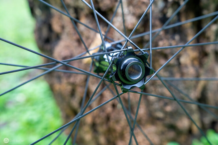 Close-up of a bicycle wheel hub with spokes radiating outward, set against a blurred natural background. The hub features a black and silver design, showcasing the details of the connection points where the spokes meet.