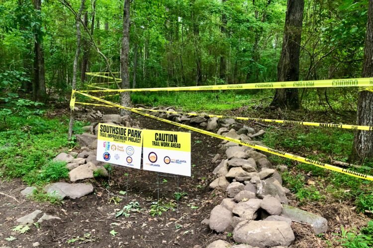 A wooded area with green foliage, featuring a sign that reads "Southside Park Trail Improvement Project" surrounded by caution tape. The trail is marked as a work zone, with stones lining the path and trees in the background.