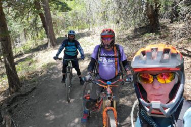 Three mountain bikers pose for a selfie on a forest trail. They are wearing helmets and protective gear. The first rider, in the foreground, has a gray beard and sunglasses, while the others are seen behind him, with one wearing a blue jersey and the other in a purple shirt. There are trees and greenery in the background, indicating a sunny day outdoors. 10k Trail mountain bike trail.