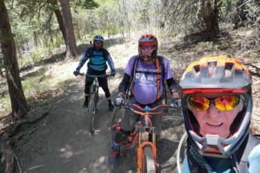 Three mountain bikers smiling on a dirt trail surrounded by trees. Each person is wearing a helmet and sunglasses, with one rider in the foreground taking a selfie. The trail is sunlit and appears to be in a natural, wooded setting. 10k Trail mountain bike trail.