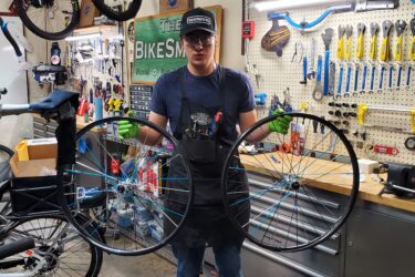 A young person in a workshop holds two bicycle wheels, showcasing their craftsmanship. The background features numerous bike repair tools and parts, with a sign reading "The Bikesmith" prominently displayed. The person is wearing an apron and gloves, indicating they are engaged in bicycle maintenance or repair. 10k Trail mountain bike trail.