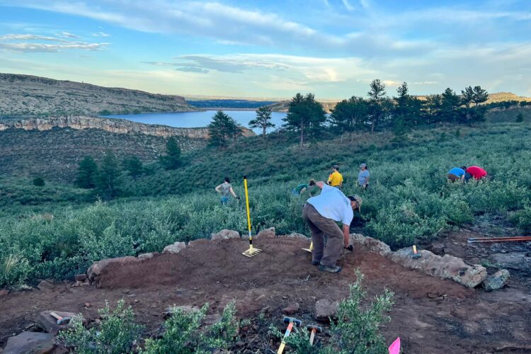 A group of people is working on an archaeological excavation site on a hillside, surrounded by greenery and overlooking a lake. The scene captures several individuals engaged in digging and surveying activities, with tools and markers scattered around. In the background, there are rocky cliffs and a clear blue sky, emphasizing the natural landscape.