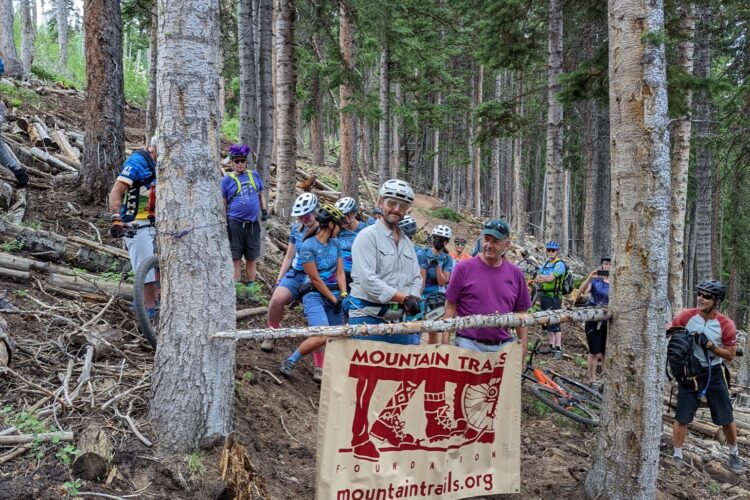 A group of mountain bikers gathered at a trail intersection in a forested area, standing near a banner that reads "Mountain Trails Foundation." The scene features several cyclists in helmets and riding gear, with trees and a dirt path visible in the background. Some cyclists are adjusting their bikes, while others are observing the area.