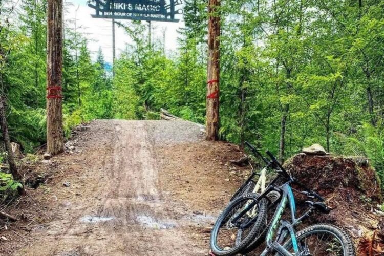A dirt trail leading through a forest, marked by a wooden archway sign reading "Niki's Magic." Two mountain bikes are parked on the side of the trail, surrounded by lush greenery and tall trees.