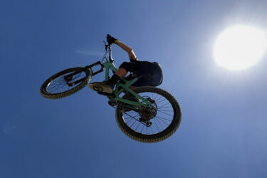 A mountain biker performing a jump against a clear blue sky, with the sun shining brightly in the background. The biker is suspended in mid-air, showcasing an action-packed moment of the sport. Erwin Trail mountain bike trail.
