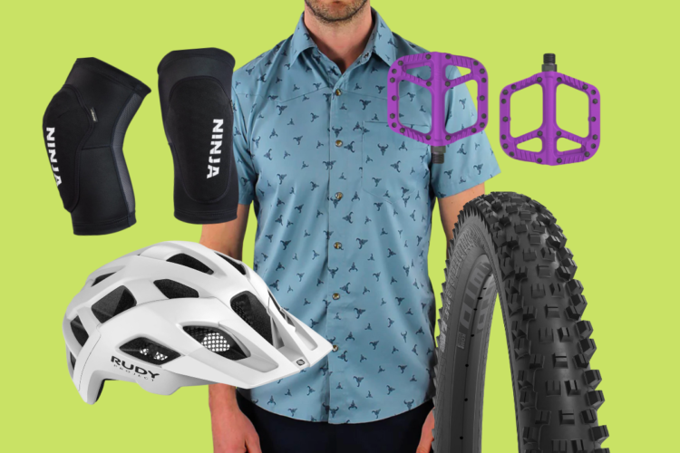 A flat lay of cycling gear and accessories against a green background. Visible items include a blue patterned shirt worn by a person, black knee pads labeled "Ninja," a white cycling helmet, purple bike pedals, and a mountain bike tire.