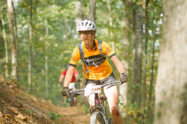A mountain biker wearing a bright orange jersey and black shorts rides along a forested trail. The rider is focused, and there's dirt on one leg, indicating a recent fall. In the background, another cyclist in a red shirt is partially visible. The scene is set amidst lush greenery and sunlight filtering through the trees. Forks Area Trail System (FATS) mountain bike trail.