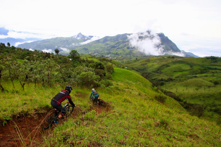Two mountain bikers navigate a winding, dirt trail through lush green hills, with a scenic mountainous landscape and misty clouds in the background. The riders, dressed in colorful biking gear, are capturing the thrill of outdoor adventure amid a vibrant, natural setting.