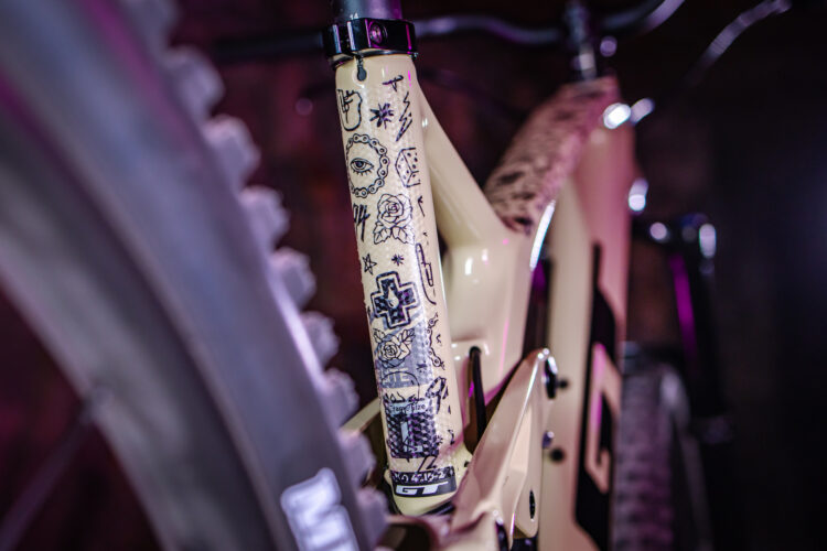 Close-up of a mountain bike frame featuring intricate black tattoo-style designs on a light-colored fork. The background includes subtle lighting effects, enhancing the bike's details and aesthetic.