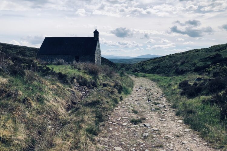 A narrow, rocky path leads through a grassy landscape towards a small, stone cottage with a sloped roof. The cottage is situated on a hillside, surrounded by rolling hills under a partly cloudy sky.