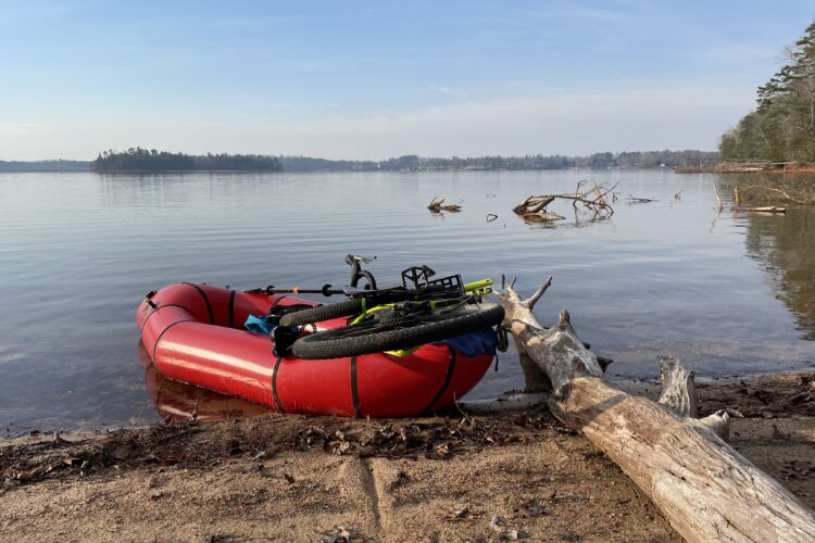 A red inflatable boat with a bicycle resting on it, positioned at the edge of a calm lake. The shoreline features sandy areas and scattered branches, with a distant island visible on the water. The sky is clear with a few clouds, suggesting a peaceful outdoor setting.