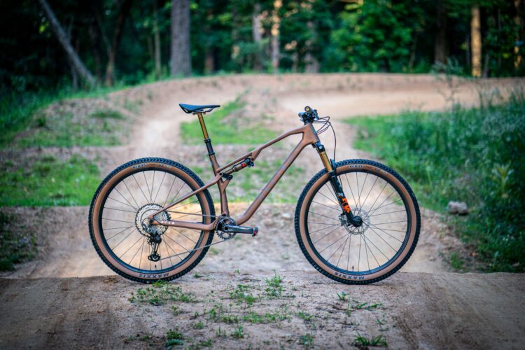 A mountain bike with a brown frame, large tires, and disc brakes is positioned on a dirt path surrounded by greenery and trees. The bike is angled to showcase its design and features, with a smooth, winding track visible in the background.