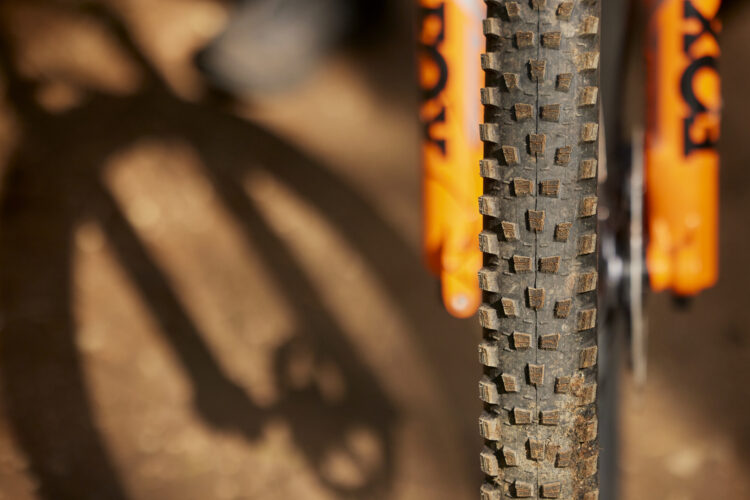Close-up of a bike tire showing its rugged tread pattern, with part of the bike's orange fork visible in the background. The ground is earthy and textured, with a shadow of the bike's components cast alongside.