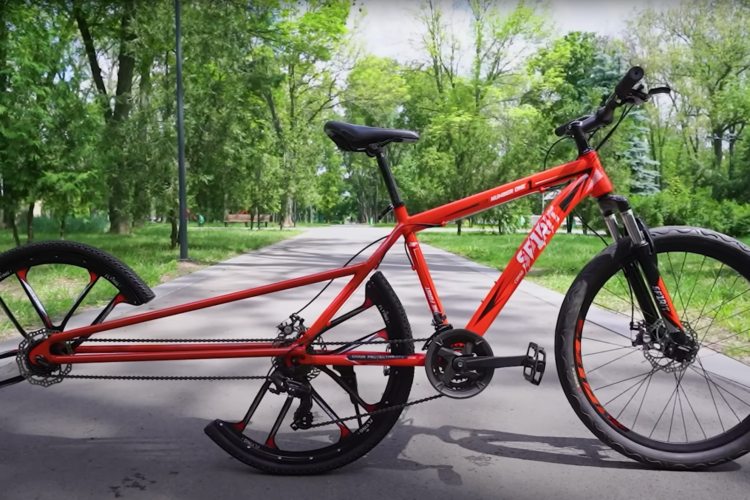 A red mountain bike is positioned on a paved path in a park, surrounded by lush green trees. The bike features a unique design with a distinctive rear wheel setup. The scene is bright and sunny, highlighting the bike's details and the natural surroundings.