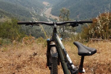 A mountain bike parked on a grassy hillside, looking out over a valley with forests and a winding river below. The landscape is partially shrouded in haze, with distant hills visible in the background. Foresthill Divide mountain bike trail.