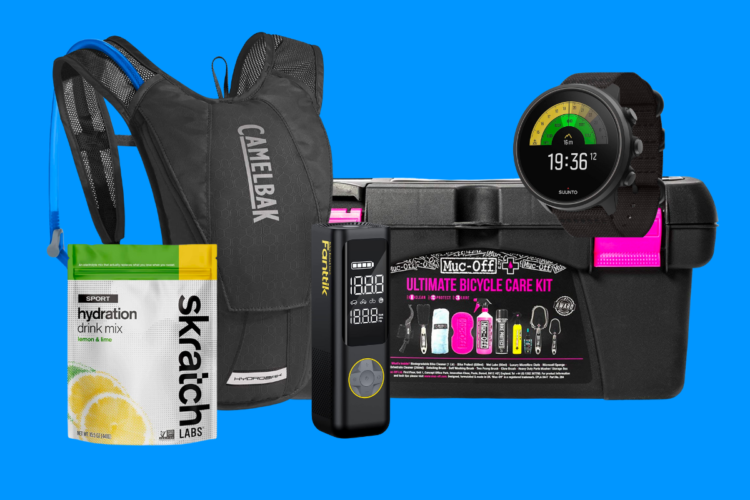 A black CamelBak hydration pack, a packet of Skratch Labs hydration drink mix (lemon and lime flavor), a Fanttik digital tire inflator, and a Suunto sports watch are displayed against a bright blue background, along with a black Ultimate Bicycle Care Kit from Muc-Off.