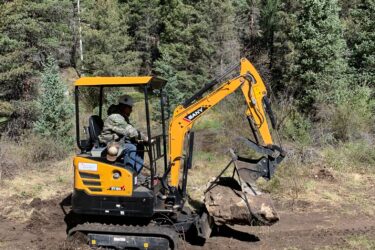 A construction worker operates a small yellow excavator in a forested area, lifting a large rock. Surrounding the machinery are green trees and sparse underbrush. The scene captures the interaction between machinery and nature in a construction project. Rito Seco Park mountain bike trail.