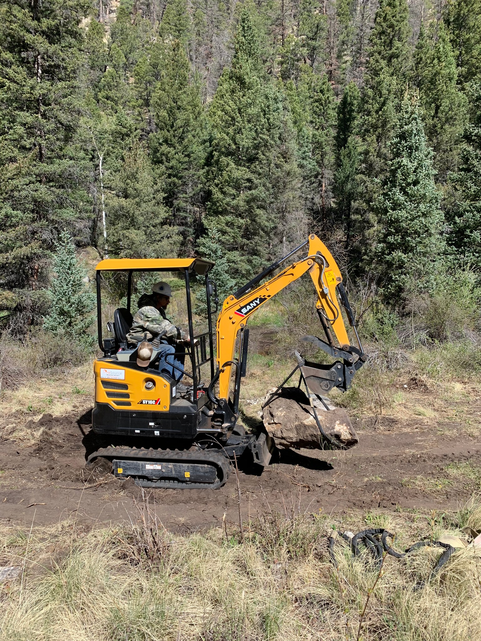 A construction worker operates a small yellow excavator in a forested area, lifting a large rock. Surrounding the machinery are green trees and sparse underbrush. The scene captures the interaction between machinery and nature in a construction project. Rito Seco Park mountain bike trail.