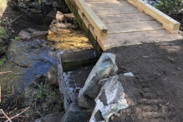 A wooden footbridge crosses over a small stream, set on a bank supported by stones and wire mesh. The surrounding area is grassy and wooded, indicating a natural trail environment. Rito Seco Park mountain bike trail.