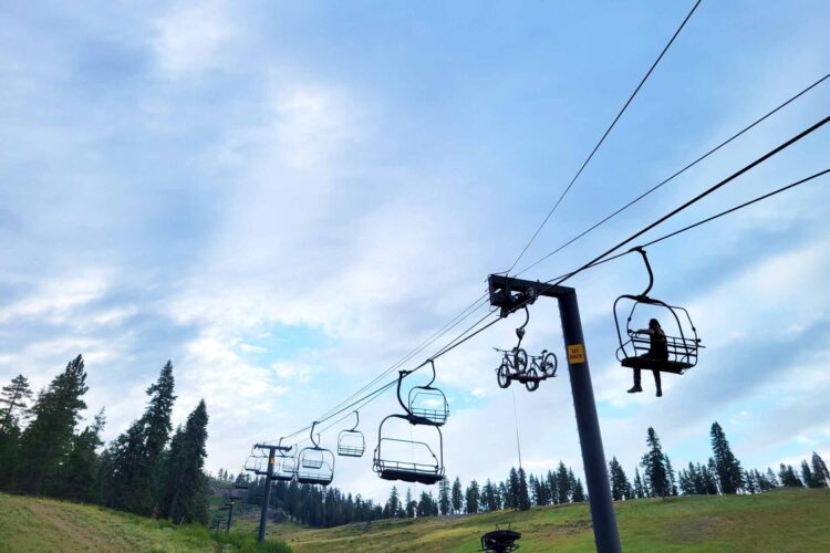 A ski lift shown against a blue sky, with a person sitting on one chair and a bicycle hanging on another. Surrounding the lift are green hills and tall trees.