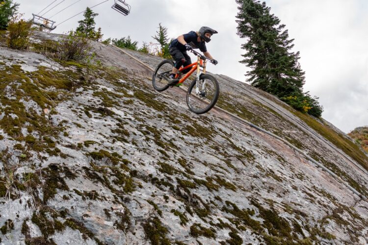 A mountain biker riding down a rocky, sloped trail covered in moss, with ski lift chairs visible in the background against a partly cloudy sky.