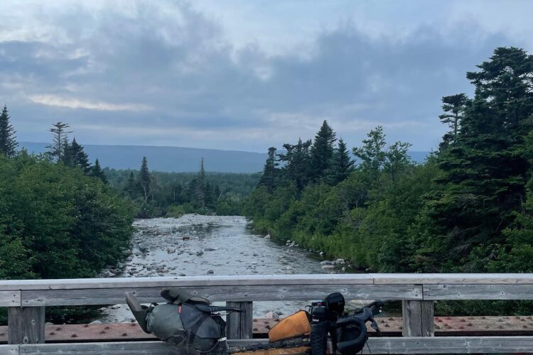 A scenic view from a wooden bridge overlooking a flowing river, surrounded by lush greenery and trees. In the foreground, a partially visible bicycle equipped for travel rests against the railing, suggesting an adventure in nature. The sky is partly cloudy, adding to the tranquil atmosphere of the landscape.