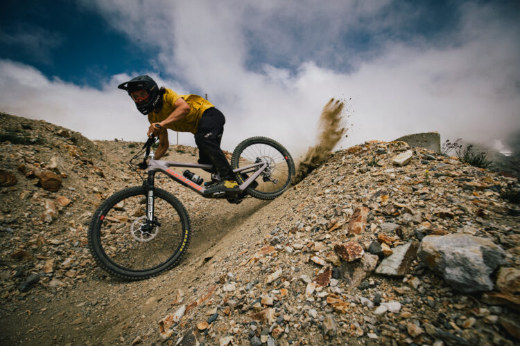 A mountain biker in a yellow shirt and black pants leans into a turn on a rocky trail, kicking up dust behind them. The bike features thick tires and appears to be navigating a challenging terrain under a partially cloudy sky.