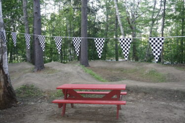 A red picnic table sits on sandy ground surrounded by trees. Above, black and white checkered flags are strung between the trees, creating a festive atmosphere. In the background, there are grassy mounds. Saranac Lake Pump Track mountain bike trail.