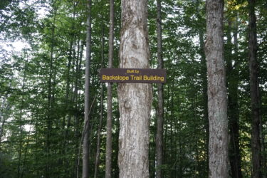 A wooden sign mounted on a tree in a forested area, reading "Built by Backslope Trail Building," with dense greenery and trees in the background. Saranac Lake Pump Track mountain bike trail.