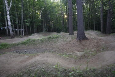 A dirt bike trail winding through a wooded area, featuring several small dirt mounds and turns. The scene is surrounded by tall, green trees, with sunlight filtering through the leaves in the background. Decorative flags are hanging from a tree, adding a festive touch to the natural setting. Saranac Lake Pump Track mountain bike trail.