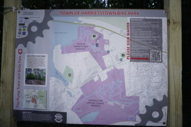 A map and informational sign for the Town of Harrietstown Bike Park, detailing the layout of bike trails and features, including the pump track and skills park. The sign includes bike park rules, safety information, and a QR code for additional resources. The map indicates surrounding areas, parking locations, and nearby landmarks, with sections highlighted in purple.  Saranac Lake Pump Track mountain bike trail.