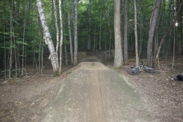 A dirt bike jump in a forested area, surrounded by tall trees and underbrush. A mountain bike is parked on the ground to the right, with a small bag nearby. The jump features a smooth, raised landing area, leading into a narrow trail that winds deeper into the woods. Saranac Lake Pump Track mountain bike trail.