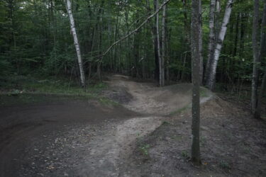 A winding dirt path through a forested area, featuring natural berms and contours, surrounded by green foliage and birch trees. The scene is set in the evening light, creating a serene atmosphere. Saranac Lake Pump Track mountain bike trail.