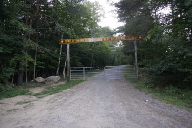 A gravel road leading to a gated entrance marked by a sign that reads "Town of Harrietstown - Elks Ball Field - Aug. 3, 1969," surrounded by trees and greenery. Saranac Lake Pump Track mountain bike trail.