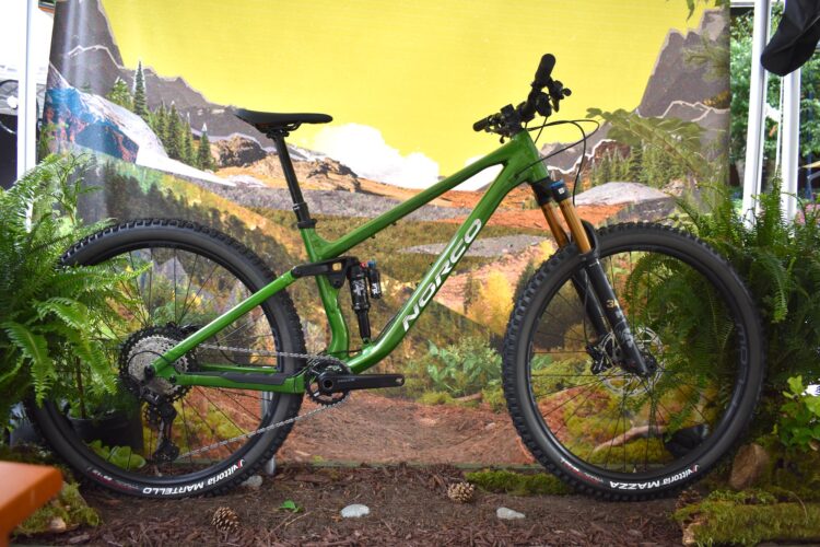 A green mountain bike displayed against a vibrant yellow and nature-themed backdrop, surrounded by ferns and foliage. The bike features a sturdy frame, black wheels, and prominent brand markings.