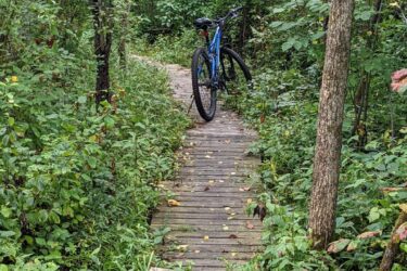 A blue mountain bike rests on a wooden path surrounded by dense greenery and trees, leading through a forested area. The scene is serene, with lush plants and leaves lining the trail. Hewen's Creek mountain bike trail.