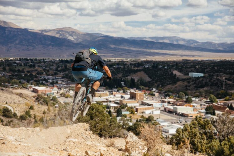 A mountain biker descending a rocky slope, with a view of a small town and mountains in the background under a partly cloudy sky.