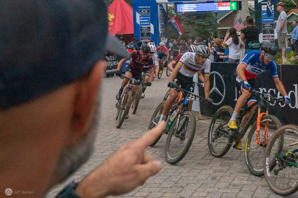 Cyclists compete in a mountain biking race, navigating a turn as they approach the finish line. A spectator is visible in the foreground, pointing towards the racers. Flags and a digital scoreboard are seen in the background, indicating the event setting.