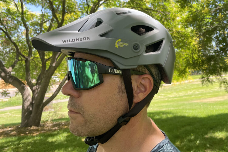 A person wearing a gray cycling helmet and mirrored sunglasses, standing outdoors with trees and grassy terrain in the background. The helmet features ventilation openings and a visor. The individual is facing to the side, showcasing the helmet and sunglasses.