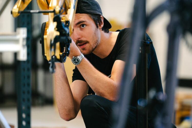 A focused individual in a black t-shirt and cap is inspecting a robotic arm in a workshop setting. The person is crouched down, looking closely at the robotics with a thoughtful expression, and a silver watch is visible on their wrist. Background elements include shelves and machinery typical of a tech or engineering workspace.