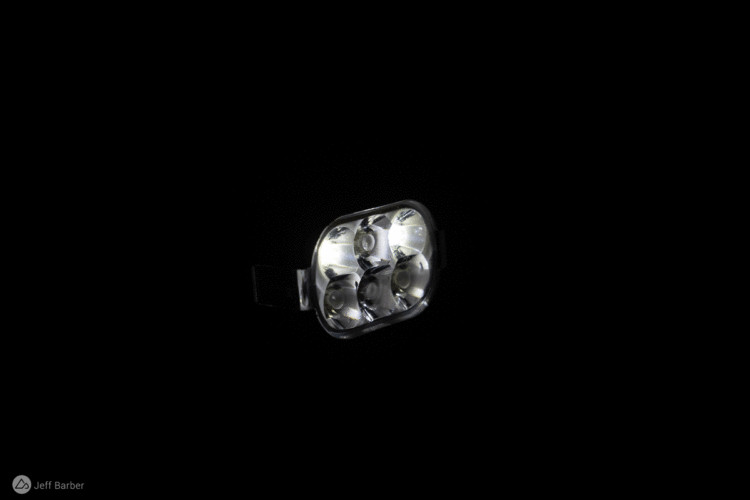 A compact, modern LED headlamp with four bright lights, illuminating against a black background.