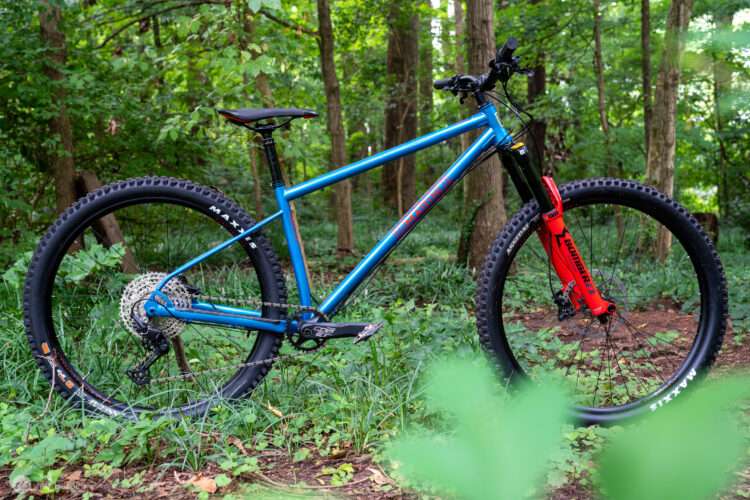 A vibrant blue mountain bike with a red fork stands in a lush green forest, surrounded by dense foliage and trees. The bike features thick tires suited for off-road terrain and is positioned on a patch of earth and grass, capturing the essence of outdoor adventure.