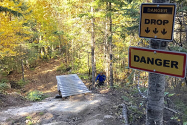 A wooded trail with a steep drop, featuring a small wooden ramp at the edge and two warning signs indicating "DANGER" and "DROP." A person in a blue jacket is visible near the bottom of the slope, surrounded by trees with autumn foliage. Leon mountain bike trail.