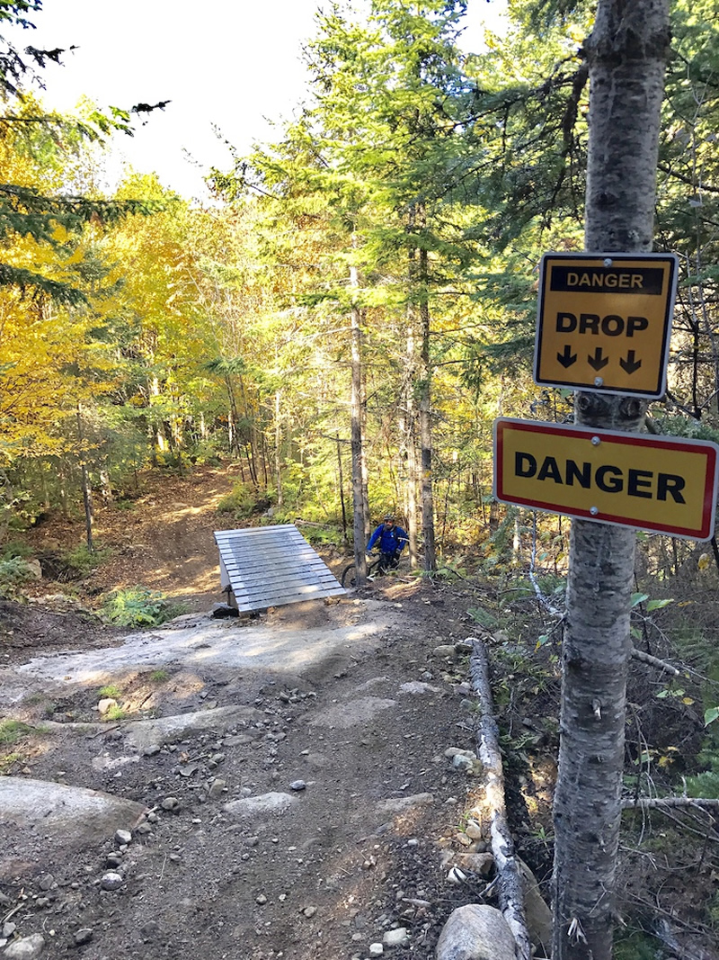A wooded trail with a steep drop, featuring a small wooden ramp at the edge and two warning signs indicating "DANGER" and "DROP." A person in a blue jacket is visible near the bottom of the slope, surrounded by trees with autumn foliage. Leon mountain bike trail.