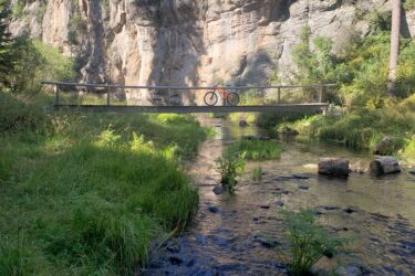 A wooden bridge spans a stream in a mountainous area, with a bright orange bicycle leaned against the bridge railing. Lush green grass and small plants surround the water, while tall trees and rugged rock formations rise in the background under a clear blue sky. East Fork mountain bike trail.