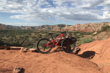 A red mountain bike rests on a rocky ledge overlooking a scenic landscape of hills and valleys under a partly cloudy sky. The vibrant red earth and greenery contrast with the rugged terrain in the background. Palo Duro Canyon mountain bike trail.