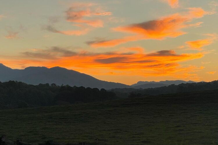 A serene landscape at sunset featuring a colorful sky with shades of orange and pink. Silhouetted mountains stretch across the horizon, while a grassy field and a wooden fence add depth to the foreground. The scene captures a peaceful rural atmosphere.