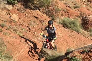 A mountain biker navigating a rocky, rugged trail surrounded by reddish dirt and sparse vegetation. The rider, wearing a helmet and gloves, appears focused as they maneuver through the terrain on a red bicycle. Palo Duro Canyon mountain bike trail.