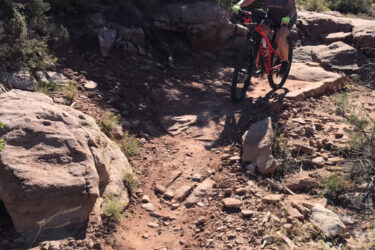 A person riding a mountain bike on a rocky trail surrounded by shrubs and trees in a natural outdoor setting. The cyclist is wearing a helmet and is positioned to navigate over a large rock, with a sunny sky in the background. Falcon Flow mountain bike trail.