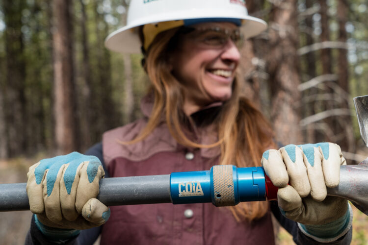 A woman in a hard hat and safety glasses smiles while holding a metal tool with her gloved hands, set against a backdrop of trees in a forested area.